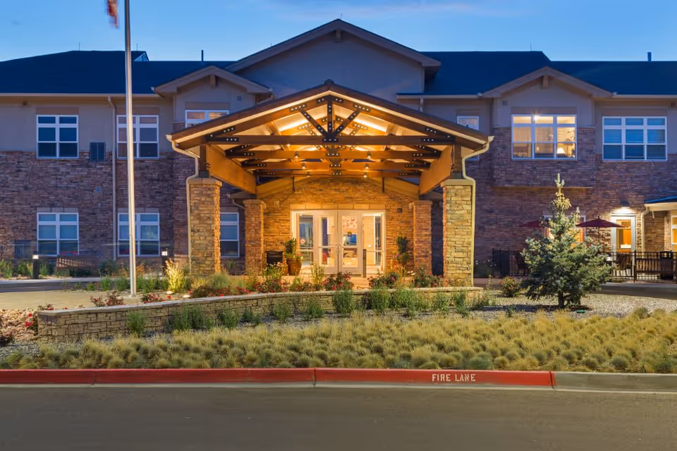 Exterior front entrance of a senior living facility named Melody Living during dusk, featuring a covered porch with warm lighting, stone pillars, landscaped garden, and multiple windows with lights on inside the building.