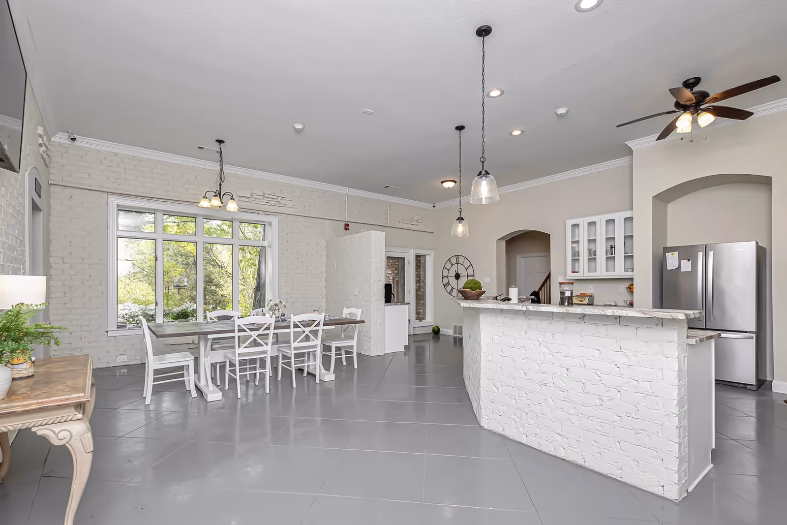 Bright and spacious kitchen and dining area with white brick walls and large windows showing greenery outside. The dining area has a wooden table with white chairs, and the kitchen features a white brick island with a marble countertop, pendant lights, ceiling fan, and stainless steel refrigerator.