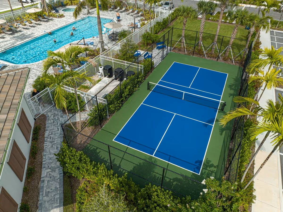 Aerial view of a fenced blue tennis/pickleball court beside a swimming pool and landscaped area with palm trees.