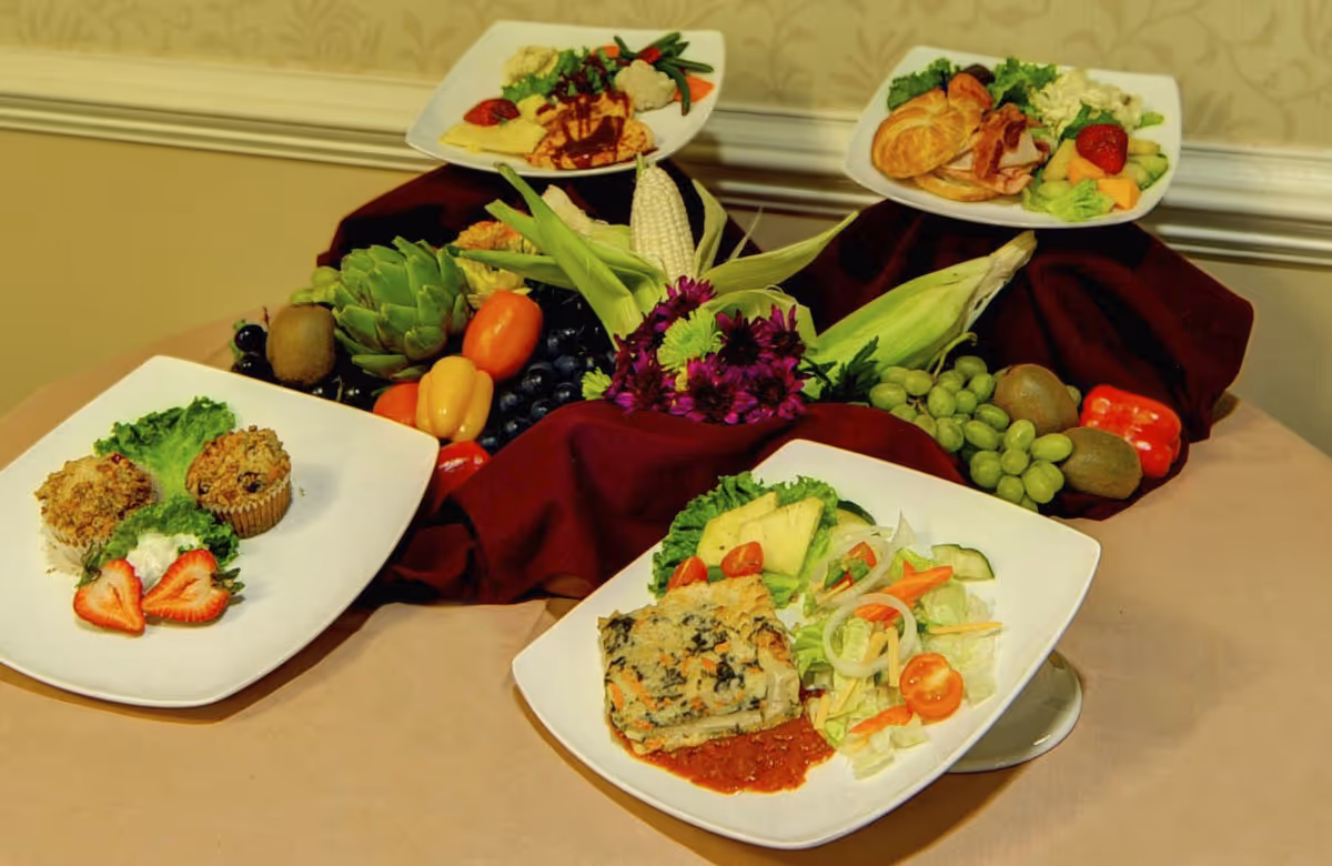 A display of four white plates with various meals including muffins with strawberries and cream, a vegetable casserole with salad, a croissant sandwich with bacon and fruit, and an omelette with vegetables. The plates are arranged on a table with a centerpiece of fresh fruits, vegetables, corn, and purple flowers on a burgundy cloth.