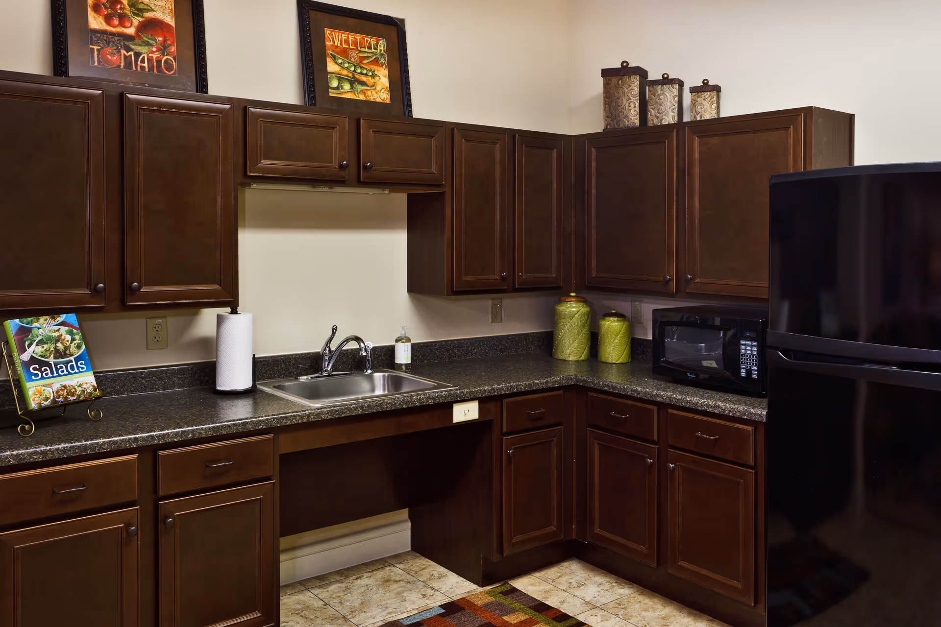 A kitchen area with dark wooden cabinets, a black countertop, a stainless steel sink with a faucet, a black microwave, and a black refrigerator. There are decorative items on top of the cabinets and framed pictures on the wall above the sink. A cookbook titled 'Salads' is displayed on the counter next to a paper towel holder.