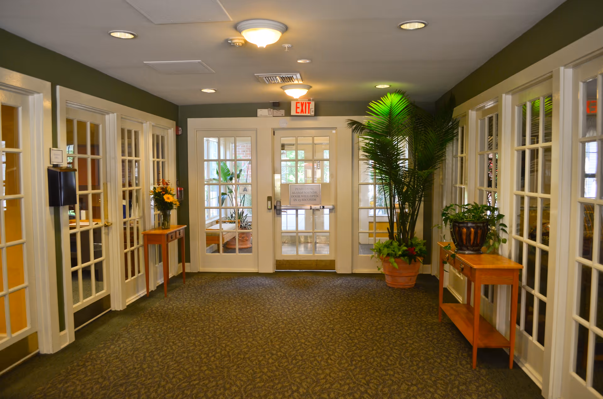 Interior hallway of a senior living facility with glass-paneled double doors at the end, flanked by potted plants and small wooden tables with decorative items. The walls are painted green and there are multiple glass-paneled doors along the sides leading to other rooms.
