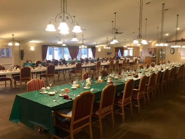 A large dining room with multiple long tables covered with green and white tablecloths, set with cups, glasses, plates, and napkins. The room is well-lit with hanging light fixtures and has several chairs arranged around the tables. Windows with curtains are visible in the background.