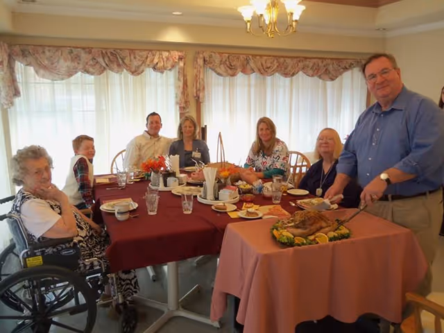 Several people seated around a dining table sharing a meal while a man carves a roast on a nearby small table.