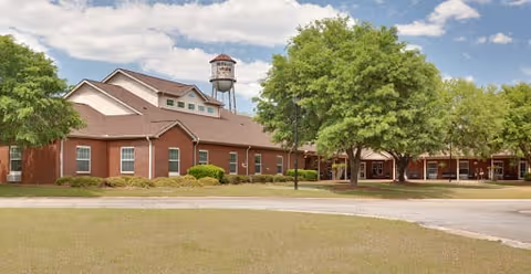 Exterior view of a single-story brick assisted living facility with a water tower in the background, surrounded by green trees and a grassy lawn under a partly cloudy sky.