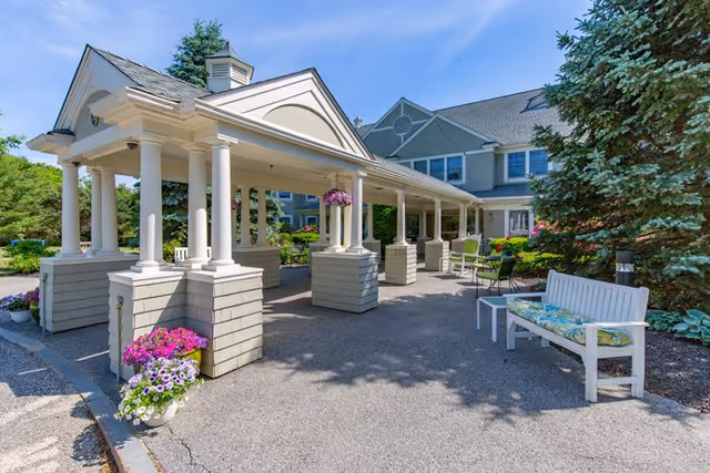 Covered portico entrance to a senior living building with columns, benches, and flowering planters.