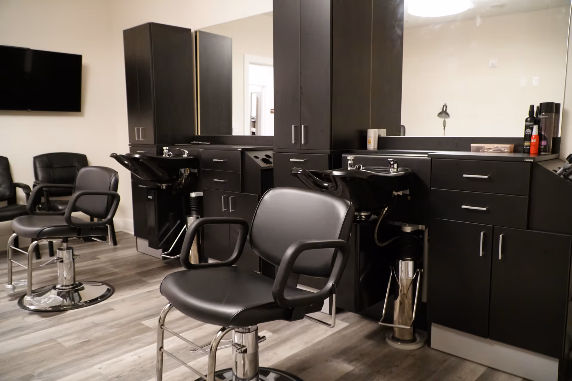 Interior view of a salon area with black salon chairs and black wash basins in front of large mirrors and dark cabinetry. The floor has a wood-like finish and there is a wall-mounted TV in the background.