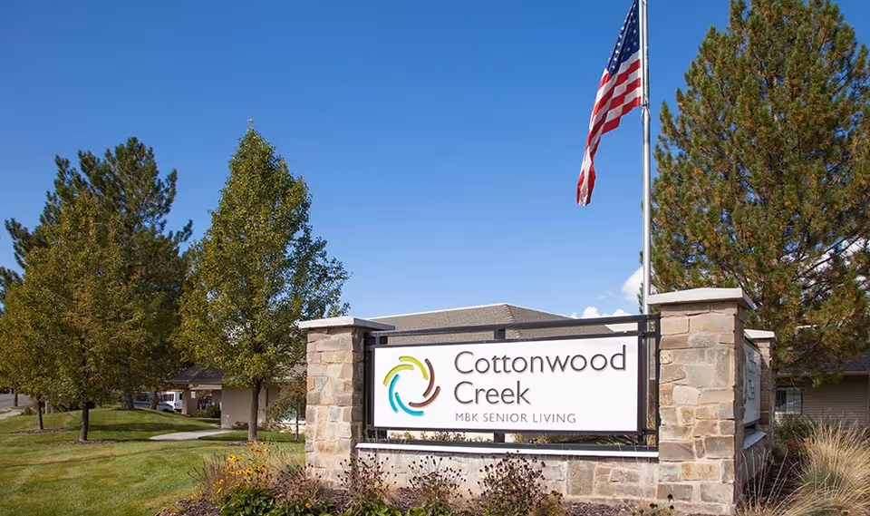 Stone entrance sign for Cottonwood Creek MBK Senior Living with an American flag, trees, and landscaped lawn under a blue sky.