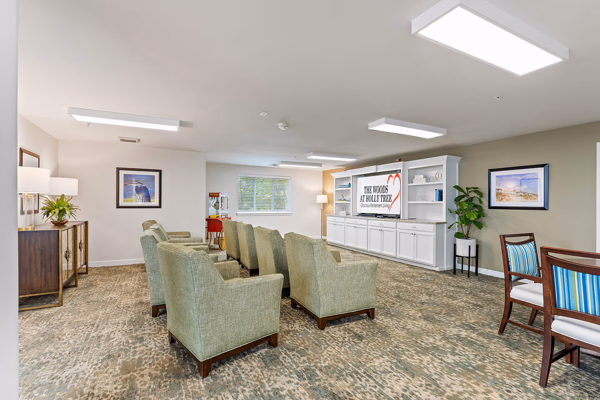 A spacious and well-lit common area in a senior living facility with rows of green upholstered armchairs facing a large white entertainment center displaying a screen with the text 'The Woods at Holly Tree Gracious Retirement Living.' The room has carpeted floors, framed artwork on the walls, a popcorn machine in the back corner, and a small table with chairs on the right side.