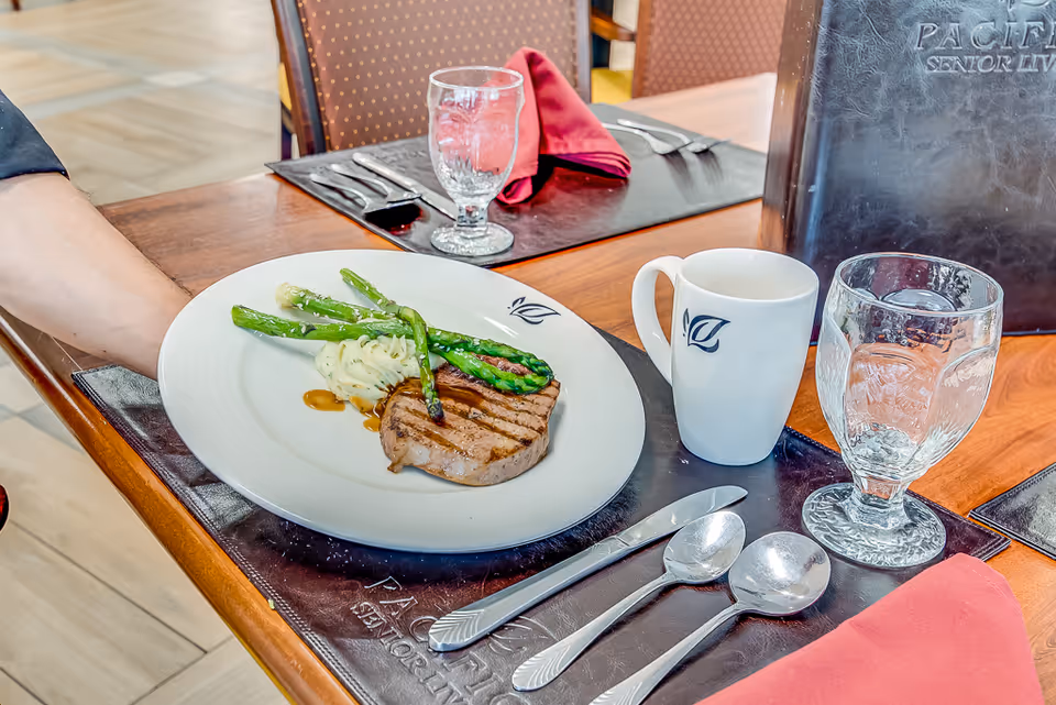 A plated meal with grilled meat, mashed potatoes, and asparagus on a white plate being served on a wooden table set with silverware, a glass of water, a white mug, and a red cloth napkin in a senior living dining area.