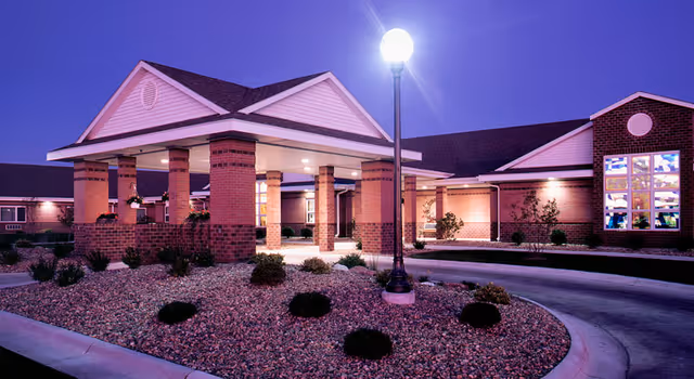 Illuminated brick entrance with a covered porte-cochère, circular driveway, and landscaped rock beds at dusk.