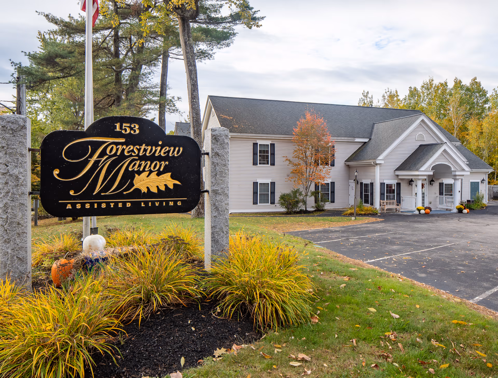 Exterior view of Forestview Manor assisted living facility with a large black and gold sign in the foreground displaying the facility name and address number 153. The building is a two-story white structure with black shutters, surrounded by trees with autumn foliage and a paved parking area.