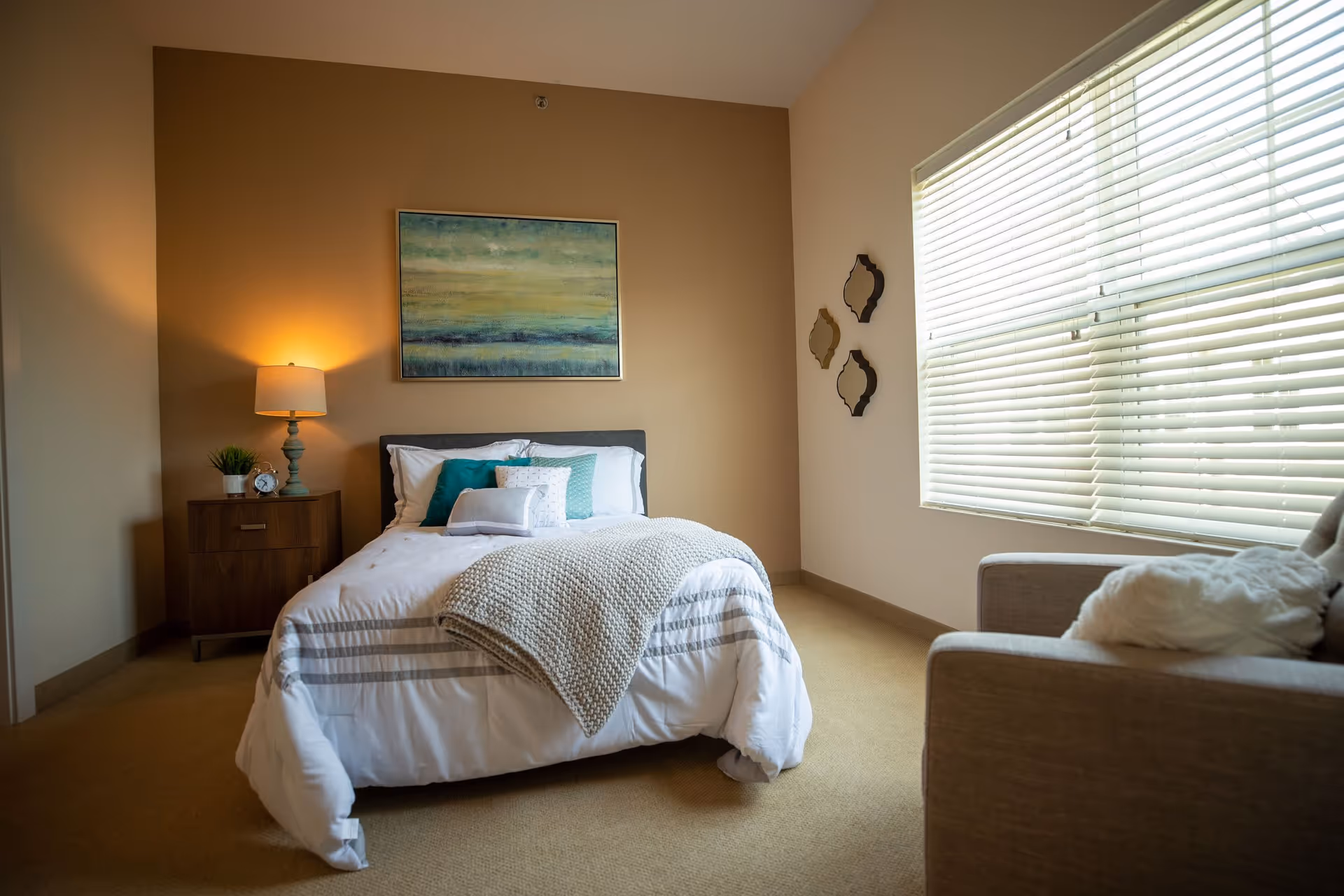 Cozy bedroom with a neatly made bed, nightstand and lamp, wall art above the headboard, and a large window with blinds.