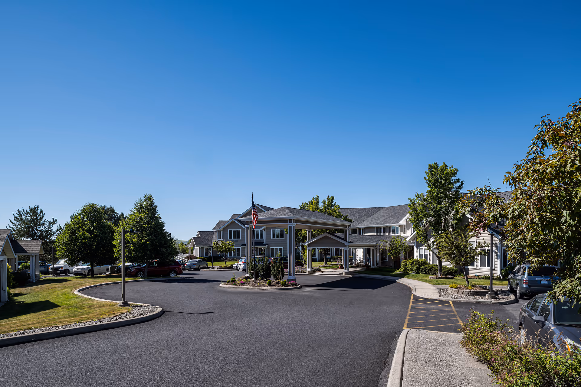Exterior view of Bishop Place Senior Living facility showing a driveway with a covered entrance, surrounded by landscaped greenery and trees under a clear blue sky.