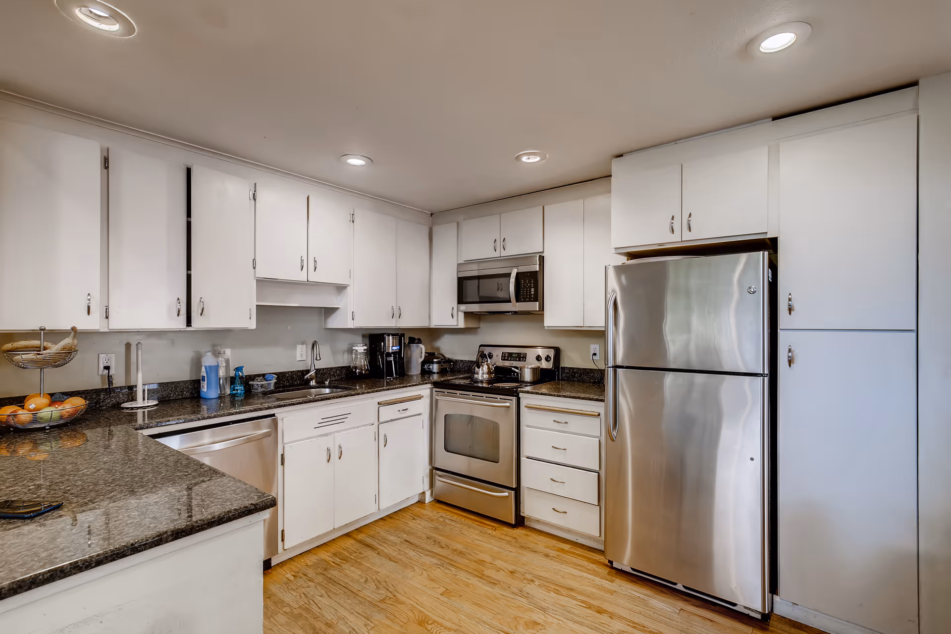 A modern kitchen with white cabinets, stainless steel appliances including a refrigerator, oven, microwave, and dishwasher. The countertops are dark granite, and the floor is light wood. There are recessed ceiling lights providing illumination.