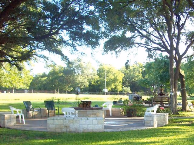 Outdoor seating area with stone walls and several chairs arranged around a central fire pit, surrounded by green grass and large trees providing shade.