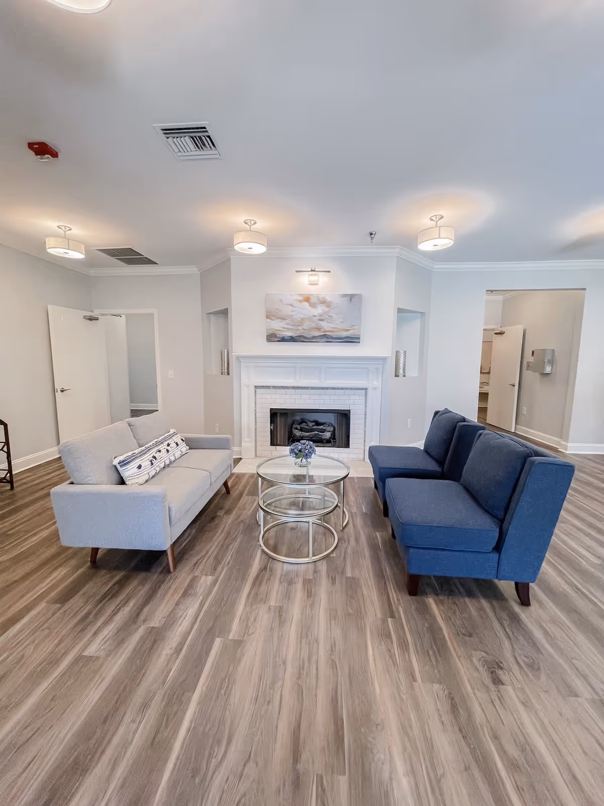 A bright and modern living room with light gray walls and wood flooring. The room features a white fireplace with a landscape painting above it. There are two sofas facing each other, one light gray and one dark blue, with a set of three round glass nesting tables in between. The ceiling has three round light fixtures, and there are open doorways visible on both sides of the fireplace.