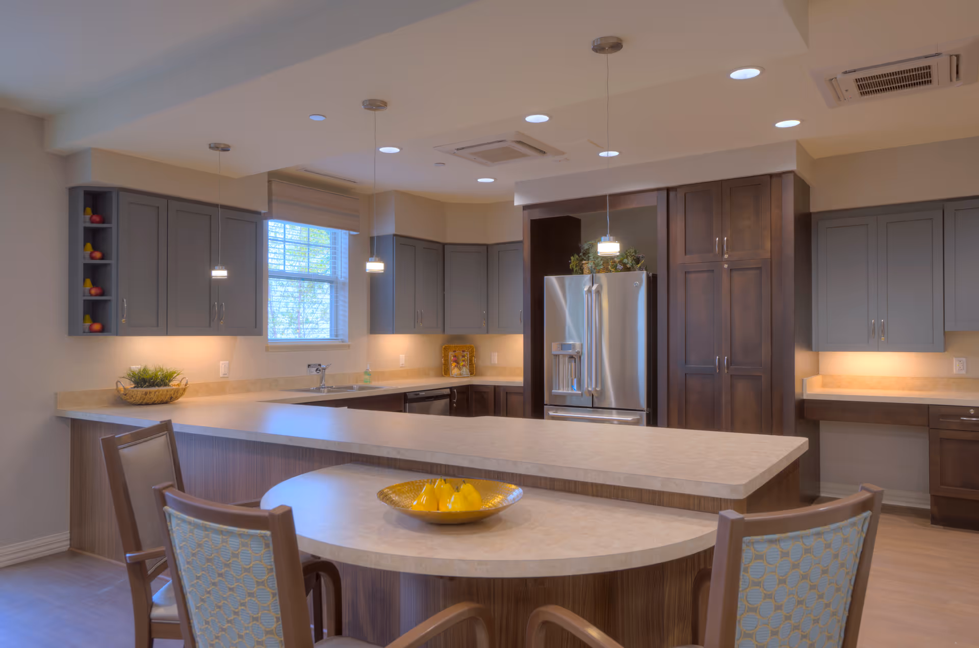 Modern communal kitchen with a large island, seating, pendant lights, stainless steel refrigerator and gray cabinetry.
