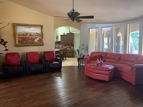 A living room with dark wood flooring, a red leather sectional sofa with a matching ottoman, and three black recliners with red cushions. A ceiling fan with lights hangs above. There is a large painting of a landscape on the wall above the recliners. In the background, an open archway leads to a kitchen area with wooden cabinets and a dining table with chairs. Large windows and a glass door let in natural light.
