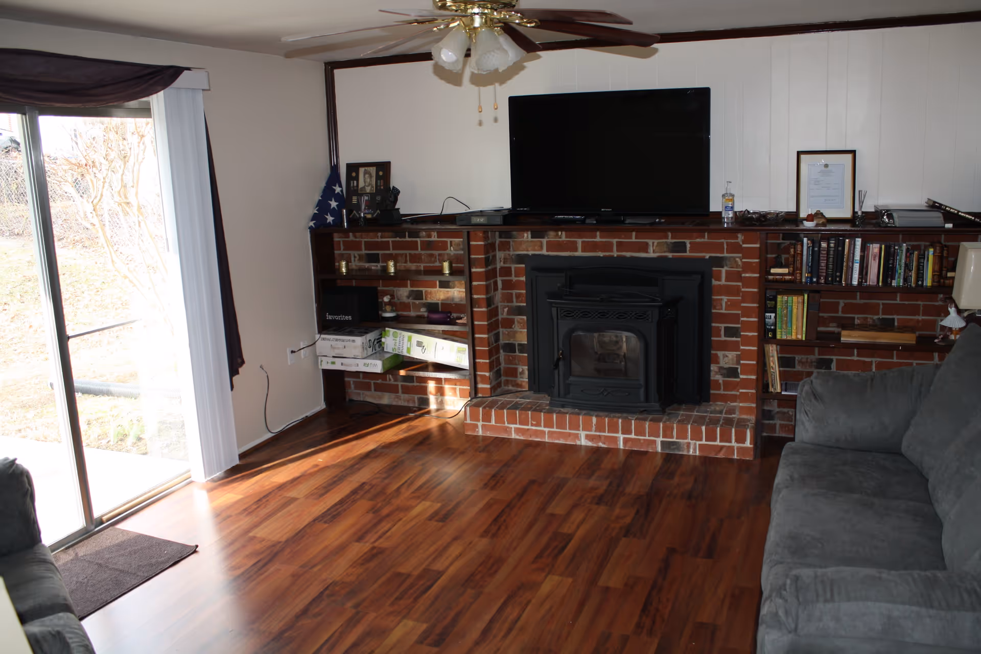 Living room with hardwood floors, a brick fireplace with built-in shelving, wall-mounted TV, sliding glass door, and a gray sofa.