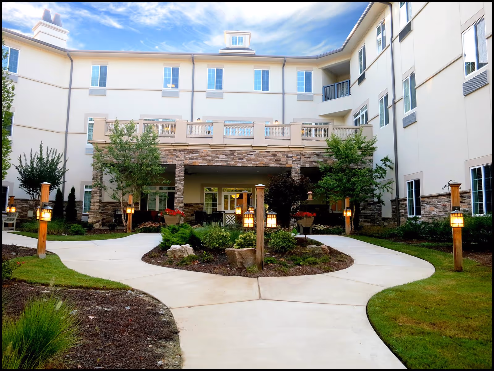 Outdoor courtyard area of a senior living facility with a paved walkway splitting into two paths around a landscaped garden with bushes and rocks. The building has three stories with multiple windows, stone accents, and a balcony above a covered patio area. Several wooden posts with lantern-style lights line the walkway.