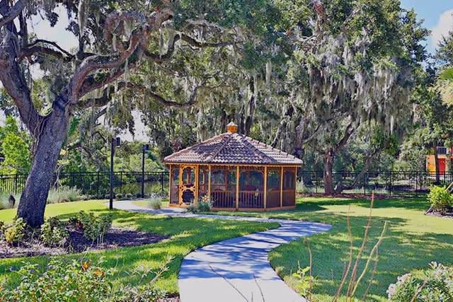 A peaceful outdoor garden area with a winding concrete pathway leading to a wooden gazebo with a tiled roof. The garden is lush with green grass, various plants, and large trees draped with Spanish moss. A black metal fence encloses the area.