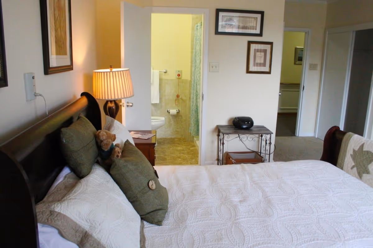 A cozy bedroom with a neatly made bed featuring white quilted bedding and green pillows, a bedside table with a lamp, and a small teddy bear. The room has light-colored walls with framed pictures. An open door reveals a bathroom with tiled floors, a toilet, and a shower with a curtain. Another doorway leads to a hallway.