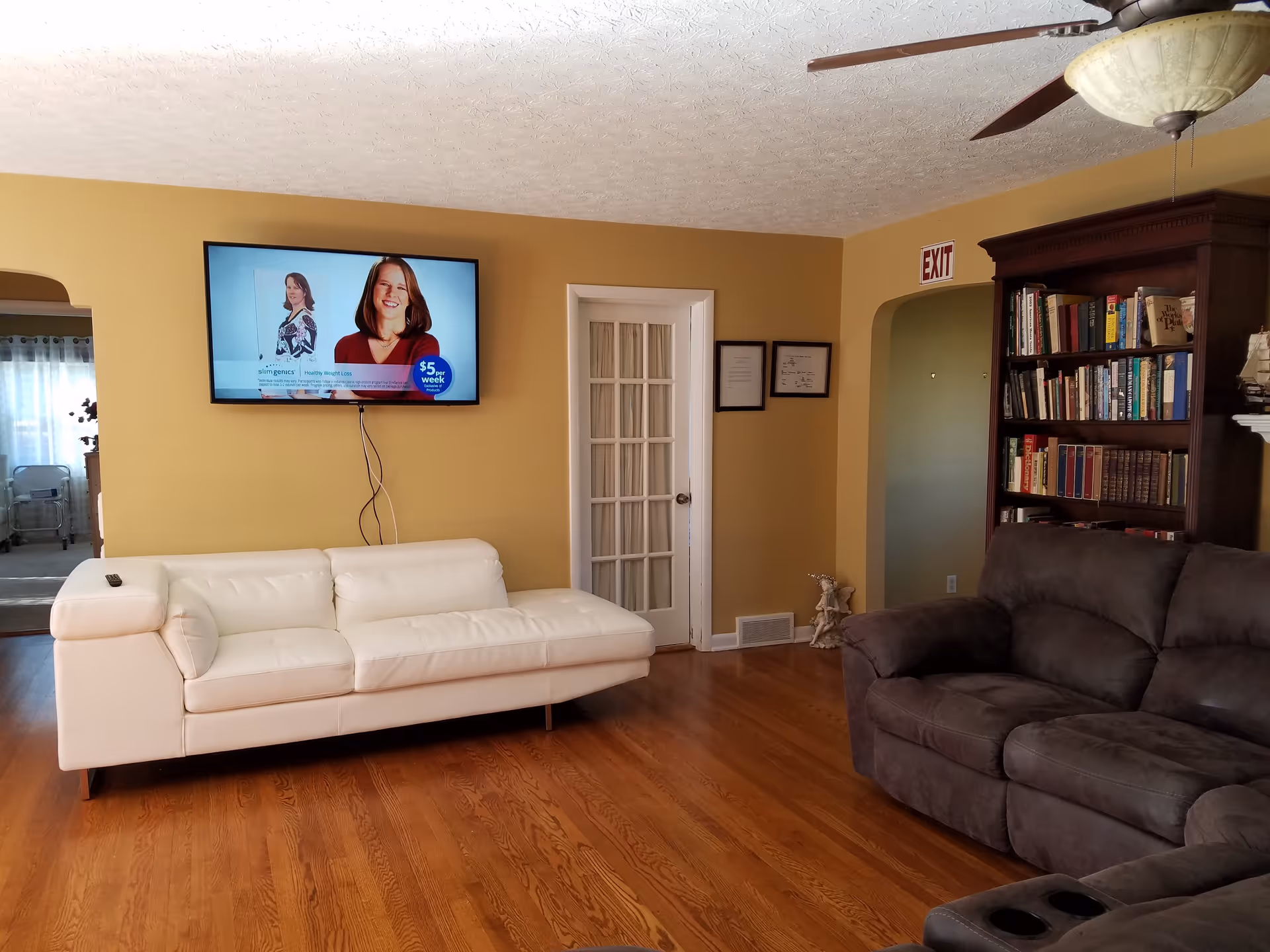 A living room with a white leather sofa and a dark gray fabric sofa. A flat-screen TV is mounted on a yellow wall above the white sofa, displaying an advertisement with a woman and text. There is a wooden bookshelf filled with books on the right side of the room. The floor is hardwood, and a ceiling fan with a light fixture is visible. A white door with glass panels and two framed certificates or pictures are on the wall. An exit sign is above an arched doorway leading to another room.