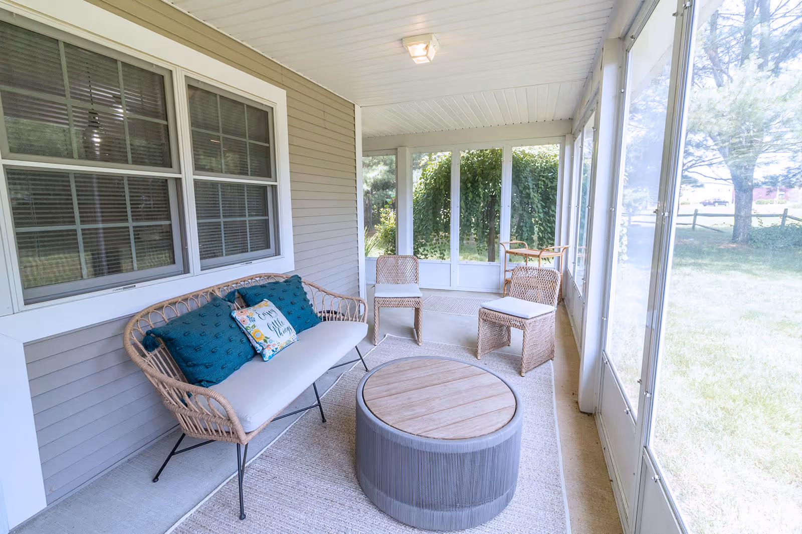 Screened-in porch with wicker seating, teal cushions, and a round wooden coffee table overlooking a grassy yard.