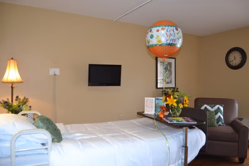 A furnished patient bedroom with a bed, wall-mounted TV, armchair, lamp, flowers and a 'Get Well Soon' balloon.