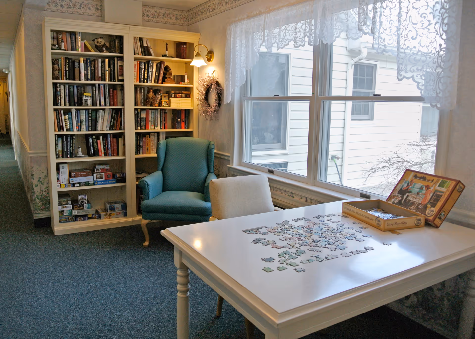 A cozy interior corner of an assisted living facility with a white table featuring an incomplete jigsaw puzzle, a blue armchair, a beige chair, a tall bookshelf filled with books and board games, and a large window with lace curtains letting in natural light.