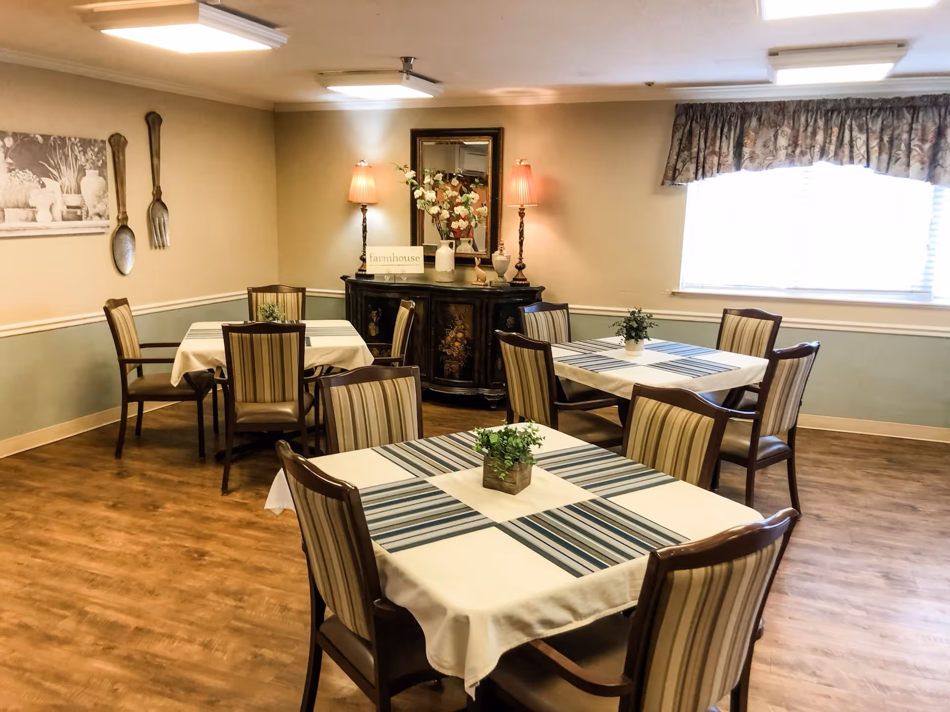 A dining room with three tables covered with white tablecloths and striped runners, each surrounded by four wooden chairs with striped cushions. The room has wooden flooring, a window with floral curtains, and a sideboard with two lamps, a mirror, and decorative items including a vase with flowers and a sign that says 'farmhouse'. On the wall, there is a black and white picture and large decorative wooden spoon and fork.