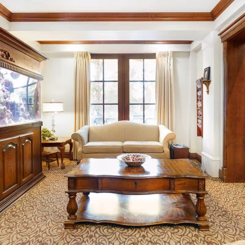 A cozy living room area with a beige sofa centered against large windows with cream-colored curtains. In front of the sofa is an ornate wooden coffee table with a decorative bowl on top. To the left, there is a large built-in wooden aquarium with colorful coral and fish. The room features patterned carpet and wooden crown molding along the ceiling edges.