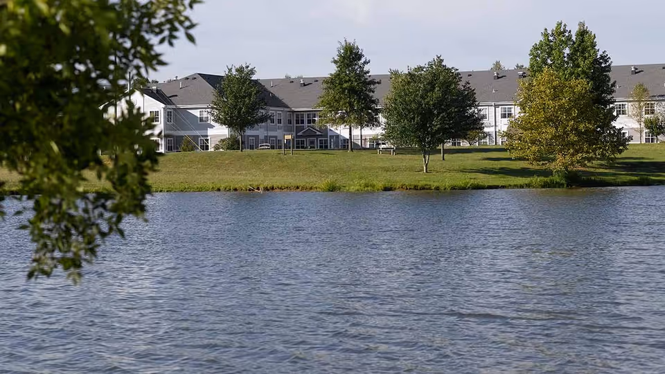 View of a large senior living facility building behind a grassy area with several trees and a pond in the foreground under a clear sky.