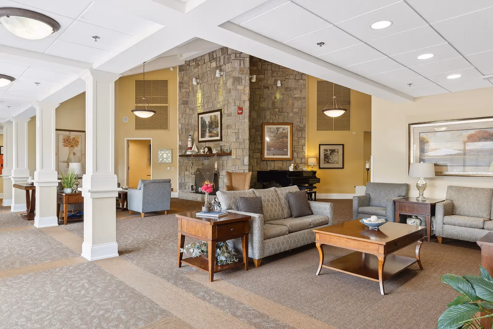 A spacious and well-lit living room area in a senior living facility featuring comfortable sofas and armchairs arranged around wooden coffee and side tables. The room has a stone fireplace with framed artwork above it, a piano in the corner, and decorative plants. The ceiling has recessed lighting and hanging light fixtures, with beige walls and carpeted floors.