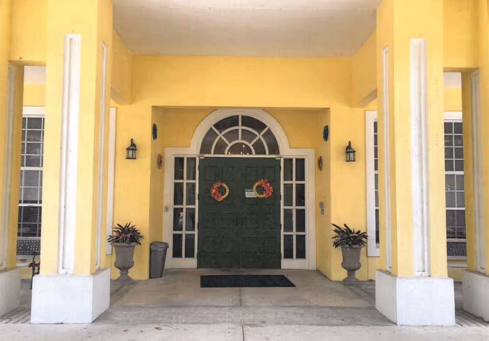 Covered entrance of a yellow building with columns and a green double door decorated with wreaths.
