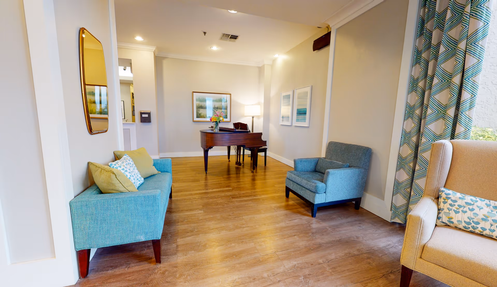 A bright and cozy living room area with wooden flooring, featuring a teal sofa with cushions on the left, a matching teal armchair on the right, and a beige armchair with a patterned cushion near a window with geometric patterned curtains. A grand piano with a vase of flowers on top is placed against the far wall, which is decorated with framed artwork and a standing lamp.