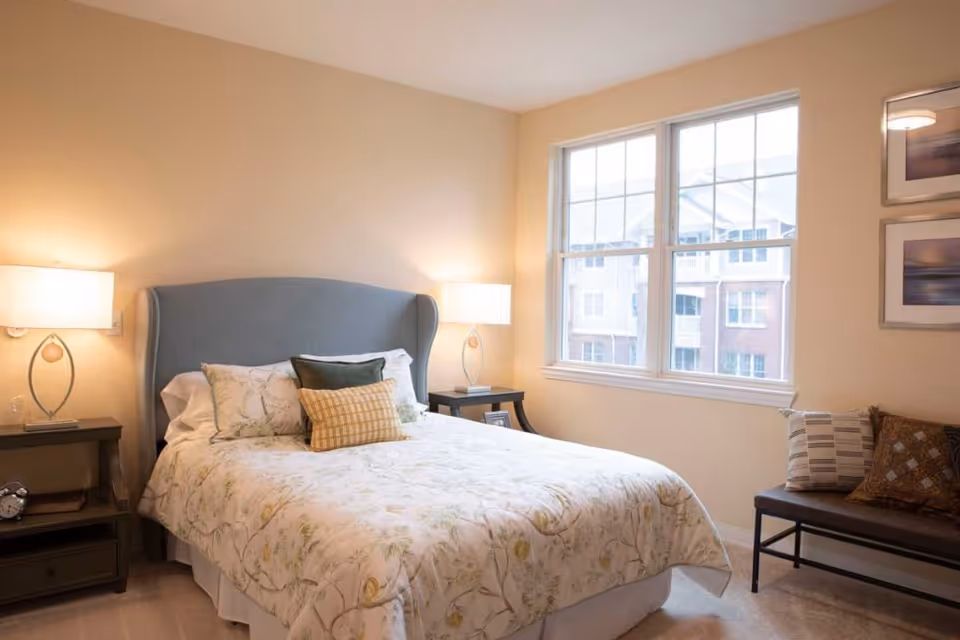 Sunlit bedroom with a queen bed featuring an upholstered headboard, two bedside tables with lamps, a large window, and a bench with pillows.