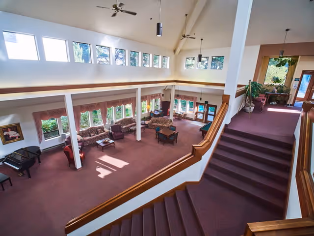 Spacious senior living facility common area with high ceilings, large windows, and natural light. The room features multiple seating arrangements including sofas, armchairs, and tables. A grand piano is positioned in one corner. Carpeted floors and a staircase with wooden railings lead to an upper level. Potted plants and framed artwork decorate the space.