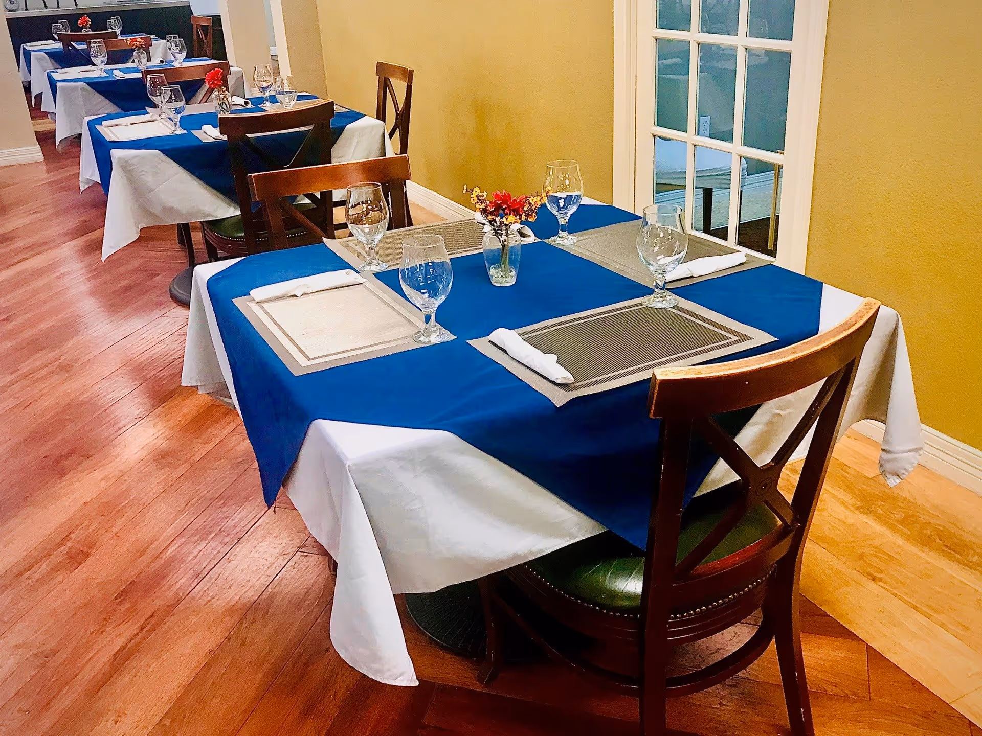 Dining area with tables covered in white and blue tablecloths, each set with placemats, napkins, water glasses, and small flower vases. Wooden chairs surround the tables, and the room has wooden flooring and yellow walls with a glass-paneled door in the background.
