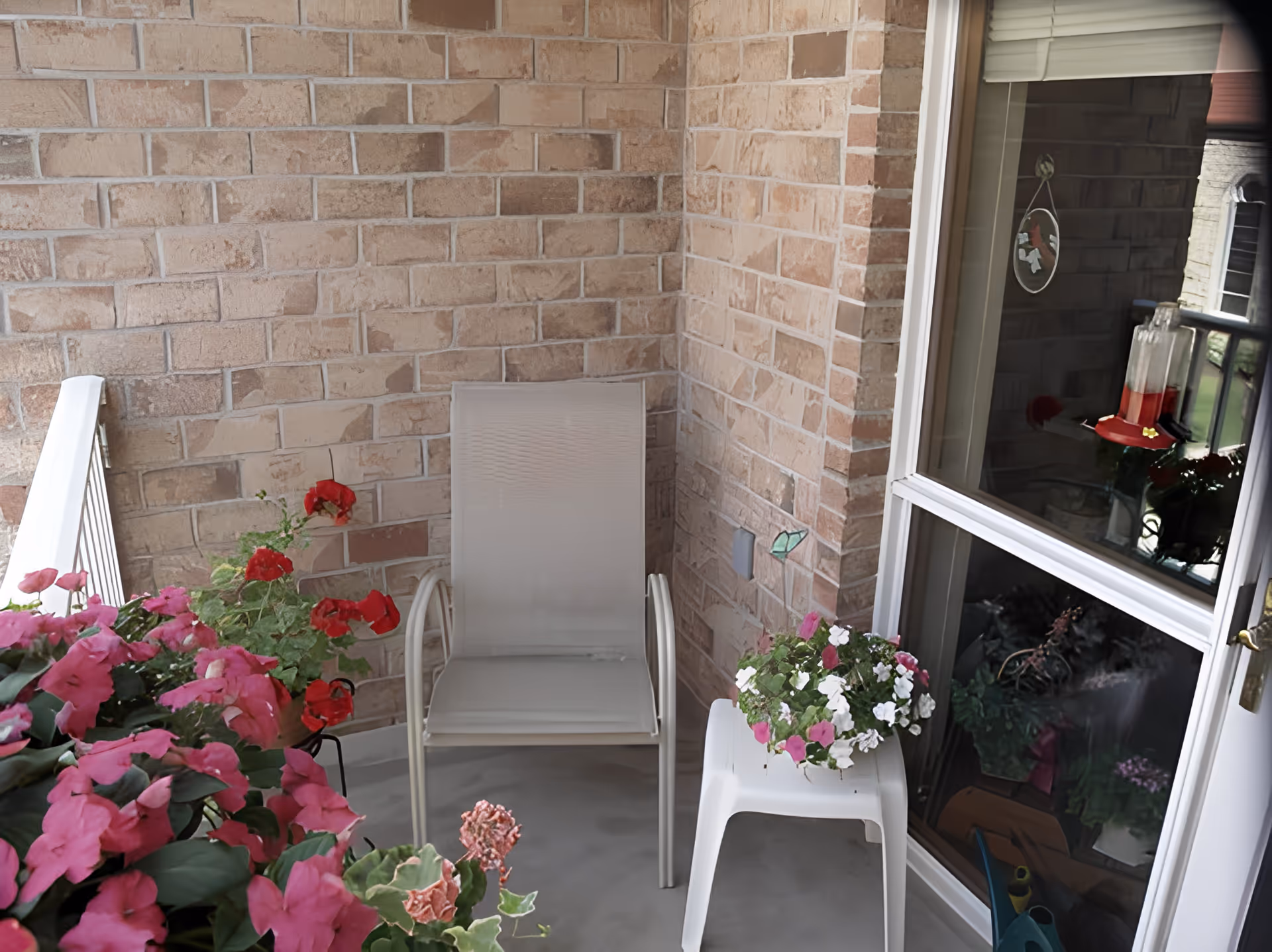 A small outdoor balcony area with a beige chair, a white side table holding a pot of pink and white flowers, and additional pink flowers in the foreground. The balcony has brick walls and a glass door with a window reflecting a hummingbird feeder and plants inside.
