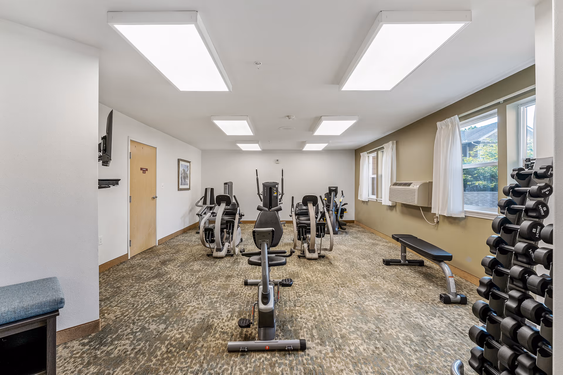 A fitness room with exercise equipment including stationary bikes, a weight bench, and a rack of dumbbells. The room has carpeted flooring, white walls with one olive green accent wall, large rectangular ceiling lights, and windows with white curtains letting in natural light.