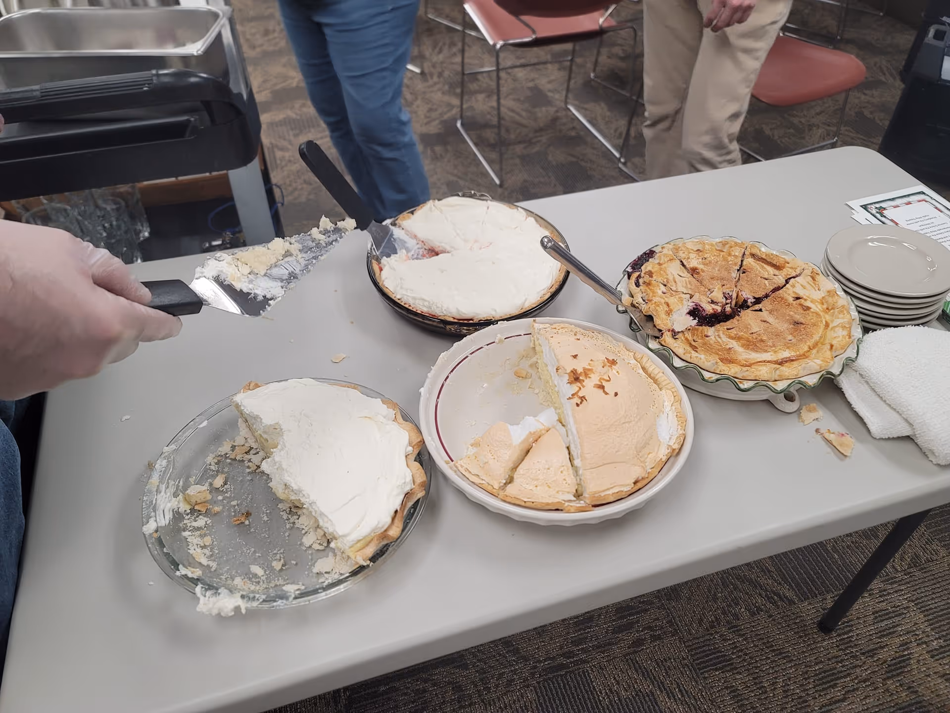 A table with four partially eaten pies, a stack of small plates, and a white towel. Two people are standing near the table, one holding a pie server with some pie crust on it. The setting appears to be indoors with carpeted flooring and chairs in the background.