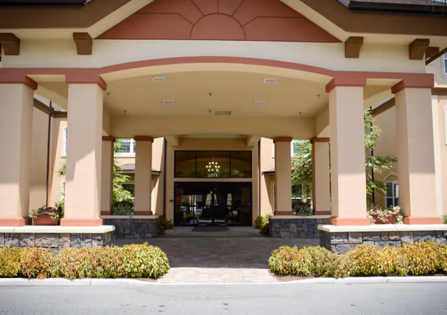 Front entrance of a building with a covered driveway supported by large columns, landscaped with bushes and plants on either side, and glass doors leading inside.