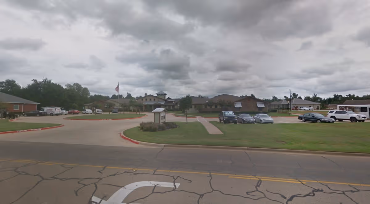 Exterior view of Spring Lake Assisted Living and Memory Care facility with a parking lot, several parked cars, an American flag on a flagpole, and a cloudy sky overhead.