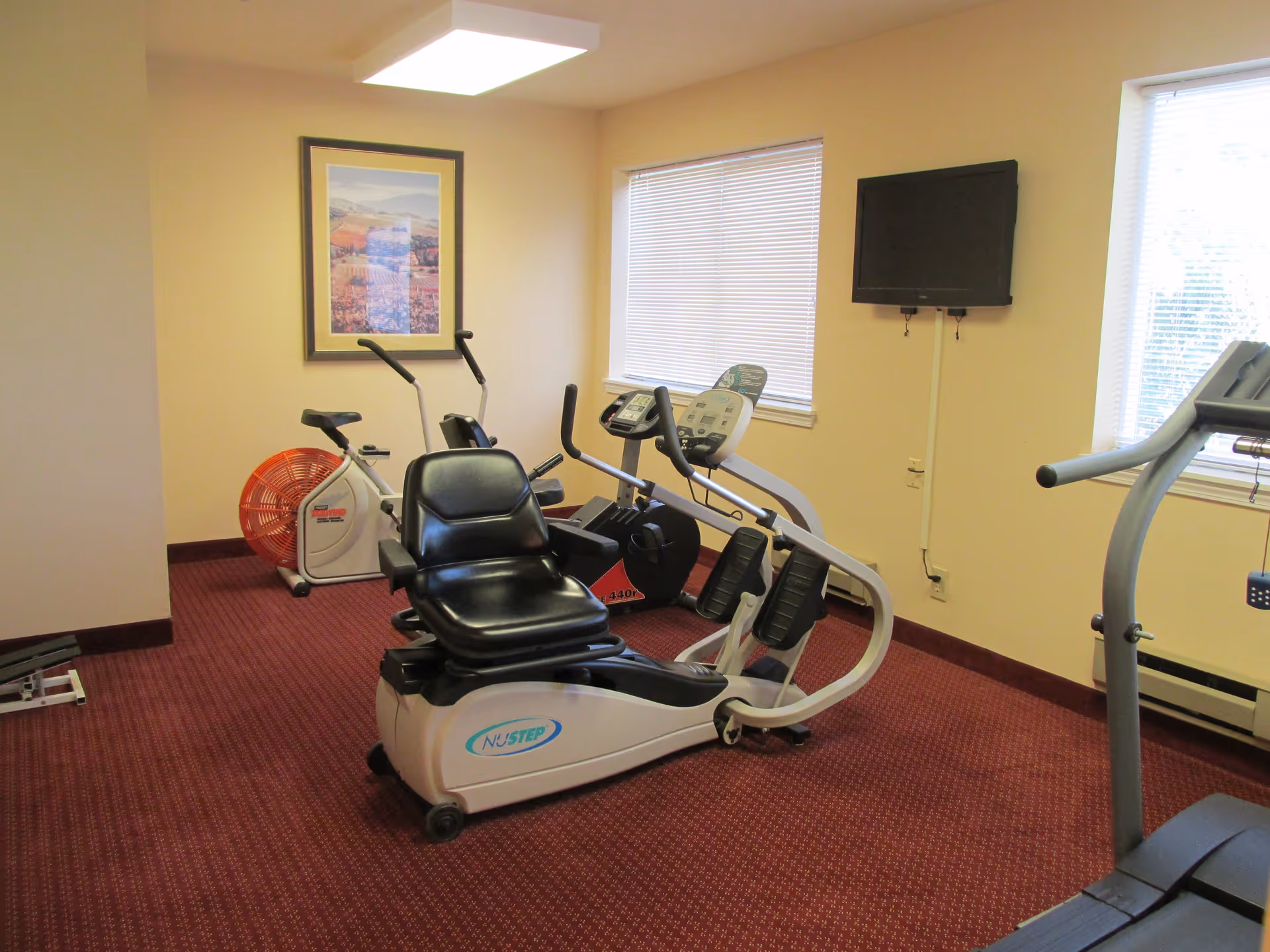 A small exercise room with a recumbent stationary bike, an upright stationary bike, and a treadmill. The room has beige walls, two windows with blinds, a wall-mounted TV, a framed picture on the wall, and red patterned carpet.
