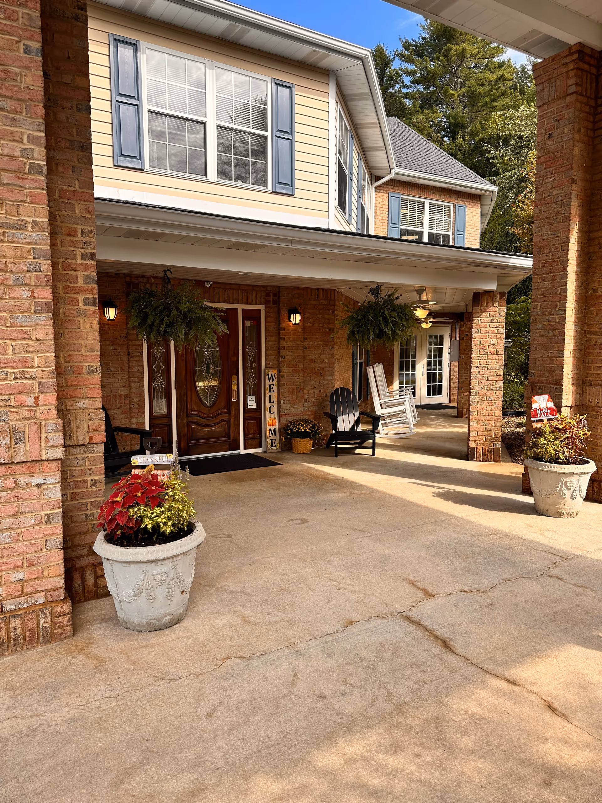 Front entrance of a senior living facility named Cedar Mountain House, featuring a covered porch with brick pillars, two large planters with colorful plants, hanging ferns, wooden rocking chairs, and a welcome sign next to a decorative wooden door with glass panels.