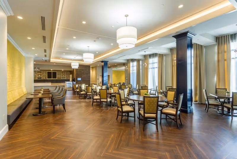 Spacious dining room with multiple round and rectangular tables surrounded by chairs with yellow upholstery. The room features large windows with beige curtains, dark wooden columns, and modern ceiling lights. The floor has a wood pattern, and there is a counter area in the background.