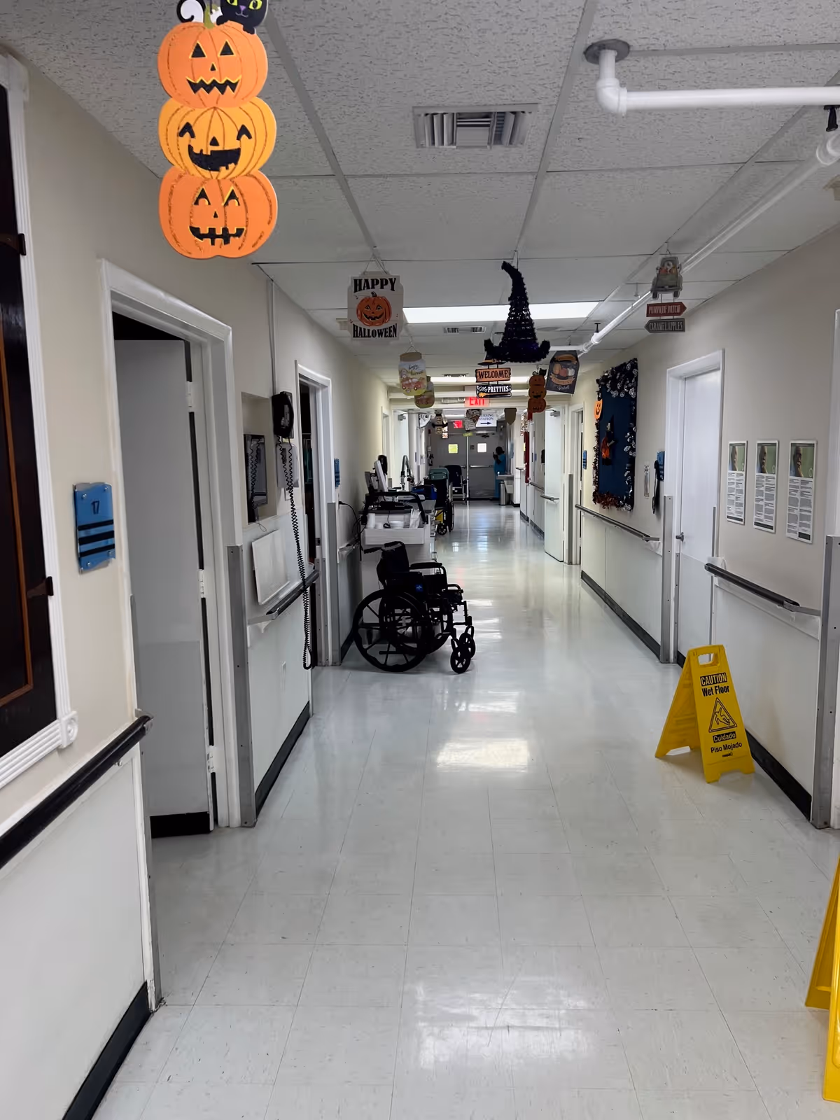 A long, clean hallway in a nursing home decorated with Halloween-themed hanging decorations including pumpkins, a witch hat, and signs. There is a wheelchair parked along the left side of the hallway and a yellow caution wet floor sign on the right side. The hallway has white walls, handrails, and several doors leading to rooms.