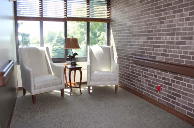 A small seating area with two beige upholstered armchairs and a wooden side table between them, which holds a lamp and a decorative horse figurine. The area has a large window with wooden blinds, a brick wall with a wooden handrail, and carpeted flooring.